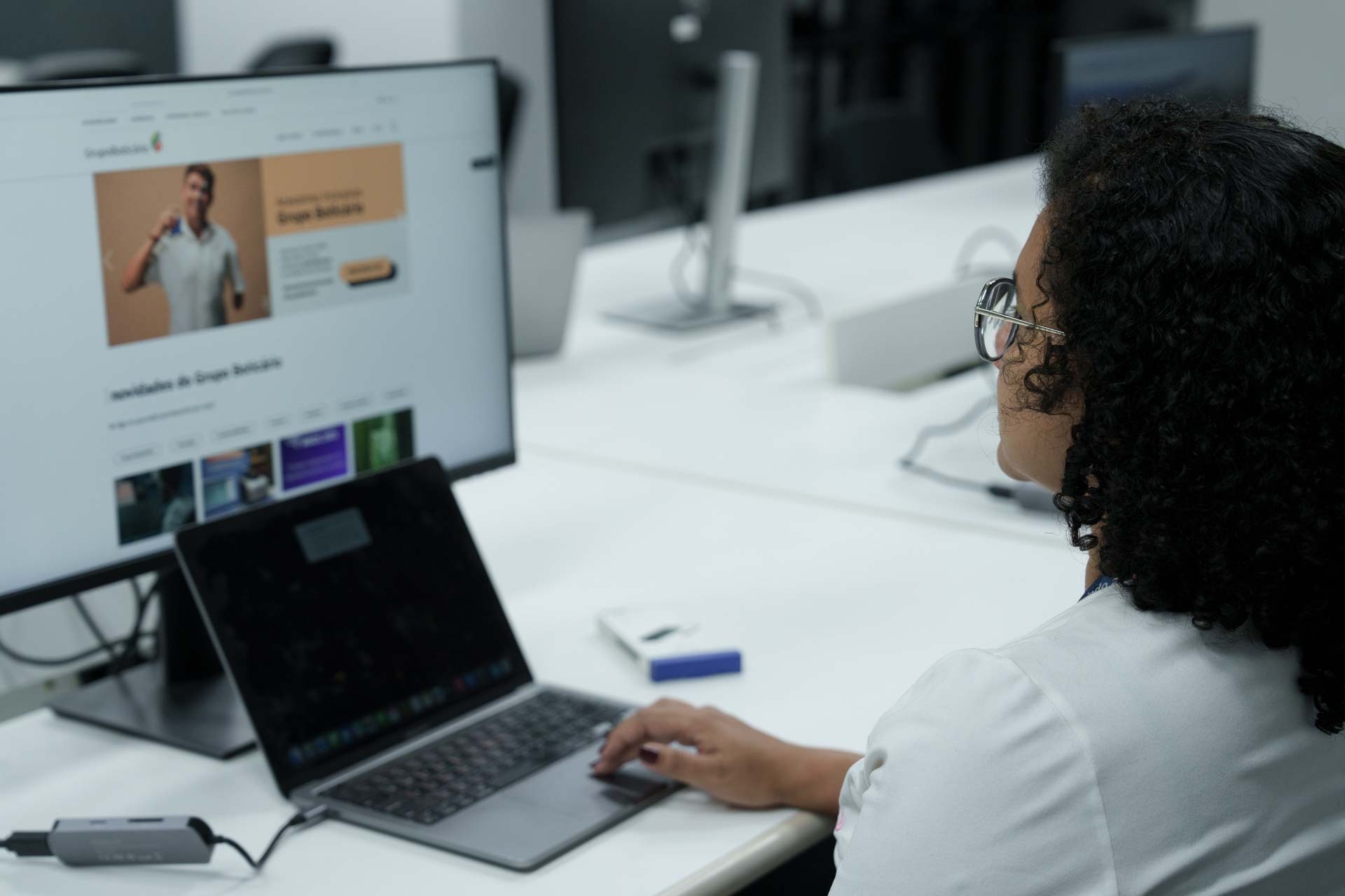 Mulher de costas, com cabelos cacheados pretos na altura dos ombros, usa óculos e blusa branca. Sentada em frente a uma mesa branca com computadores, ela utiliza um notebook.